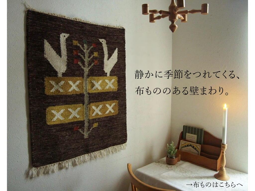 北欧ヴィンテージのタペストリーを壁に飾った部屋の一角。木製キャンドルホルダーやリネンのクロスが並ぶ、布もののある静かな壁まわりのインテリア。