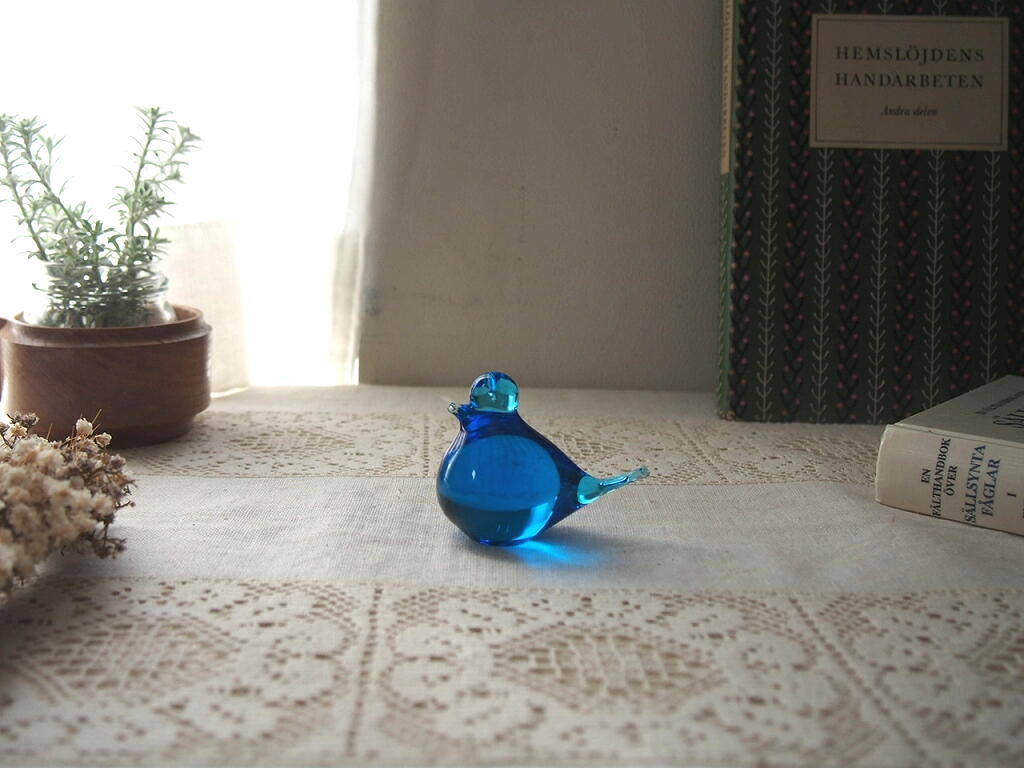 木の鉢植えや本と並んだスウェーデン製ガラスの青い小鳥。窓辺の光が透ける北欧インテリアの一角。 antenna