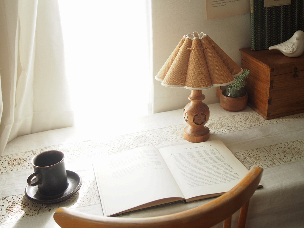 本とカップを置いたデスクをやさしく照らす木製テーブルランプのある風景。 antenna